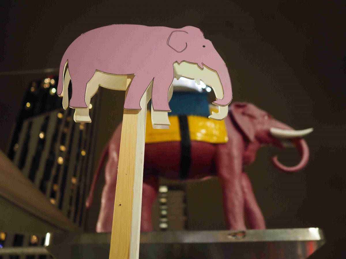 A pink elephant miniature is displayed in the background while a flat pink elephant drawing on a stick is in focus